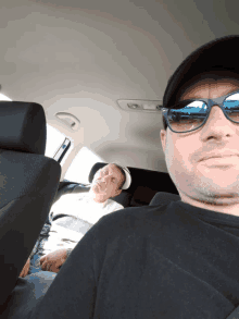 a man wearing sunglasses sits in the back seat of a car with another man sleeping in the back seat