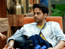 a man in a denim jacket sits in an orange chair with his legs crossed