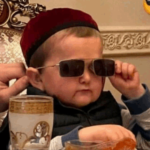 a little boy wearing sunglasses and a red hat is sitting at a table with a glass of water .