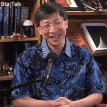 a man in a blue shirt is smiling in front of a microphone and a book called orbital paths on a shelf