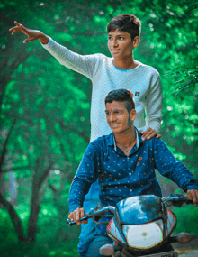 two young men are posing for a picture one of them is riding a motorcycle and the other is standing behind him with his hand up