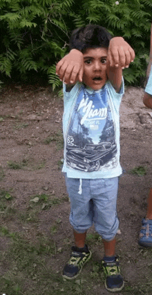 a young boy wearing a blue shirt with a car on it is standing in the grass with his hands behind his head .