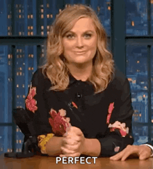 a woman is sitting at a table with her hands folded in front of her and the word perfect is on the screen .