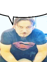 a man with a beard wearing a superman shirt is sitting down with a speech bubble above his head .