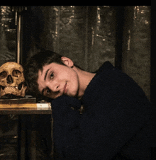 a young man in a blue sweater leans his head on his arm next to a skull