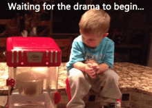 a little boy sits in front of a popcorn machine with the words " waiting for the drama to begin "