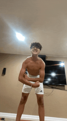 a shirtless young man wearing white adidas shorts is flexing his muscles