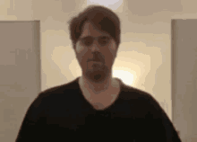 a man with a beard is wearing a black shirt and standing in a room with a light on .