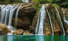 a waterfall in the middle of a forest is surrounded by rocks and trees and is surrounded by a body of water .