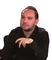 a man with a beard is sitting at a table with his hands folded in front of him