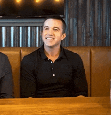 a man in a black shirt is smiling while sitting at a table in a restaurant .