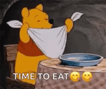 winnie the pooh is sitting at a table with a towel around his neck and a plate of food .