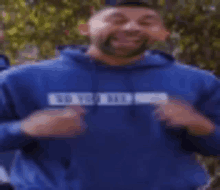 a man with a beard wearing a blue hoodie is standing in front of a tree .