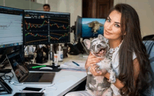 a woman sitting at a desk holding a dog in front of two computer monitors