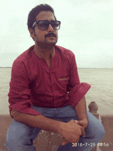 a man wearing sunglasses and a red shirt is sitting in front of a body of water on a date of 2018-7-25