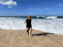 a man in a black shirt stands on the beach giving a thumbs up