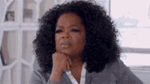 oprah winfrey is sitting in front of a window with her hand on her chin and making a funny face .