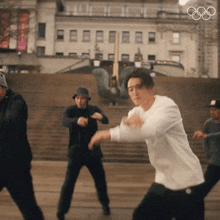 a man in a white shirt is dancing in front of a building with the olympic rings in the background