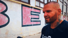 a man with a tattoo on his neck stands in front of a wall with the letter b painted on it