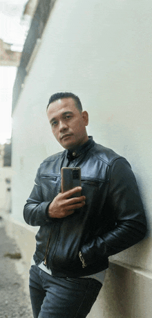 a man in a black leather jacket leans against a wall while holding a cell phone