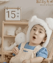 a little boy wearing a bear hat is sitting in front of a calendar that says feb 15