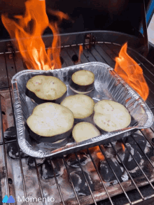 an aluminum foil pan filled with sliced eggplant is cooking on a grill with a momento logo in the corner