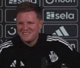 a man wearing a black and white adidas shirt smiles in front of a sign that says sela