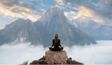 a woman is sitting in a lotus position on top of a mountain surrounded by clouds .