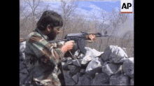 a man in a camouflage uniform is holding a rifle in front of a stone wall with an ap logo in the corner