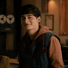 a young man wearing a vest and a hoodie smiles in front of two record players