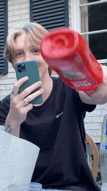 a man is holding a bottle of ketchup and taking a picture with his phone