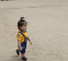 a little girl wearing overalls and a yellow shirt is walking on the beach