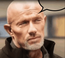 a bald man with a gray beard looks at the camera with a speech bubble above his head