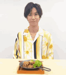 a man in a yellow and white striped shirt sits at a table with a pan of food on it