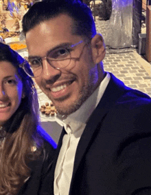 a man in a tuxedo and glasses smiles next to a woman in a white shirt