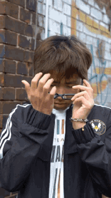 a man wearing a black jacket and a white shirt with the word team on it adjusts his glasses in front of a brick wall