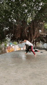 a man in a white shirt and black pants is doing a trick on a concrete floor under a tree