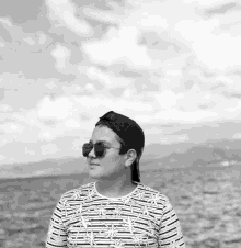 a boy wearing sunglasses and a baseball cap stands in front of the ocean