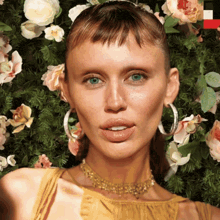 a woman wearing hoop earrings and a gold necklace stands in front of a wall of flowers