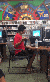 a man in a red shirt is sitting at a desk in front of a wall with a painting of a sun