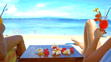 two people sit at a table on a beach holding drinks