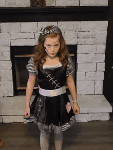 a little girl in a black and white dress stands in front of a brick fireplace