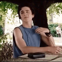 a young man in a blue tank top is sitting at a table under a tree with a pen in his hand .