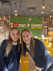 two girls pose in front of a wall that says i heart mac and cheese and more