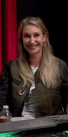 a woman in a green jacket is smiling while sitting at a desk with a bottle of water on it .