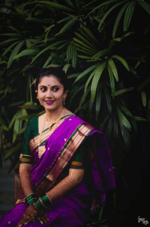 a woman wearing a purple saree and a green top is sitting in front of a tree