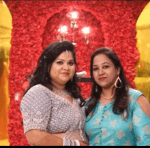 two women are posing for a picture together in front of a wall of flowers .