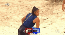 a woman in a blue shirt stands on a sandy beach with a scoreboard behind her that says ' gunduler '