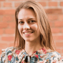 a woman with long blonde hair wearing a floral shirt smiles in front of a brick wall