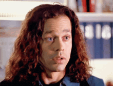 a man with long curly hair looks surprised in front of a bookshelf